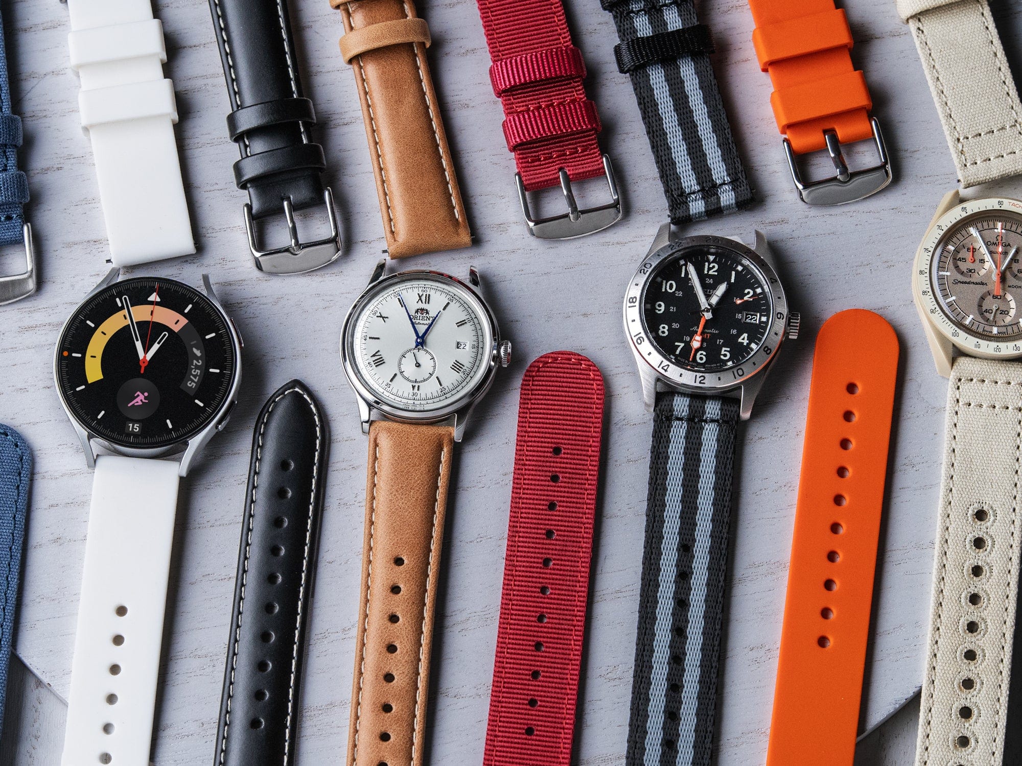 Flat lay of multiple watches with leather, nylon, and silicone straps in various colors on a light surface