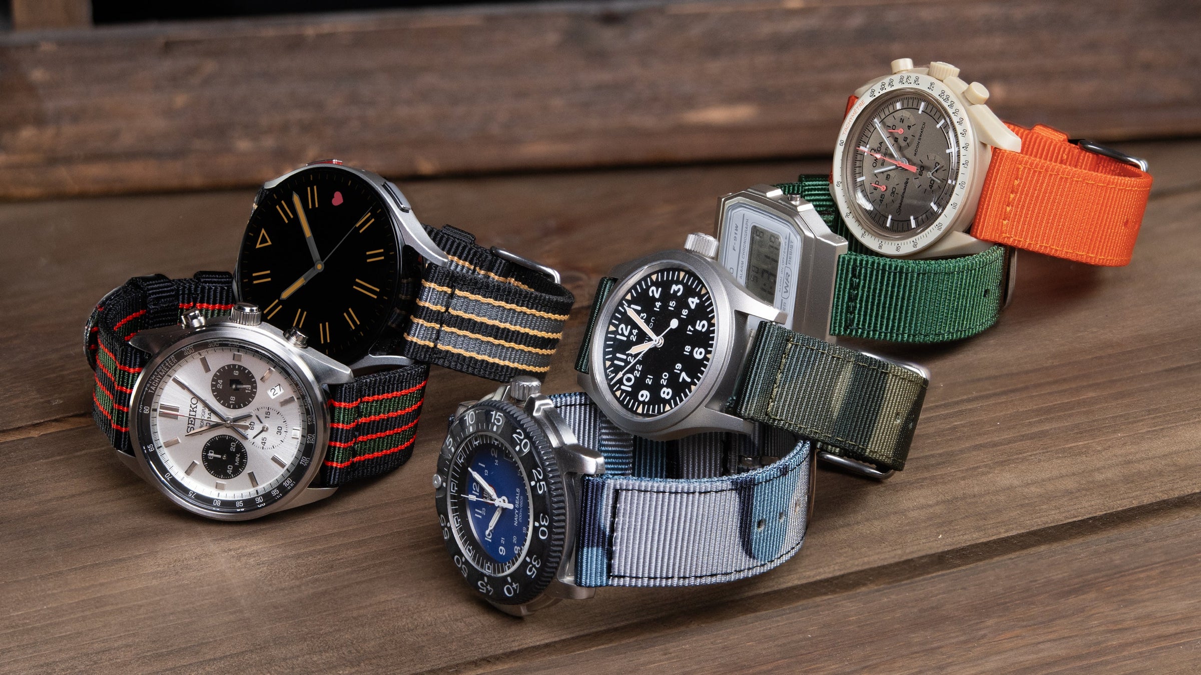 Five watches with new nylon straps in striped, camo, orange, and green displayed on a wood surface