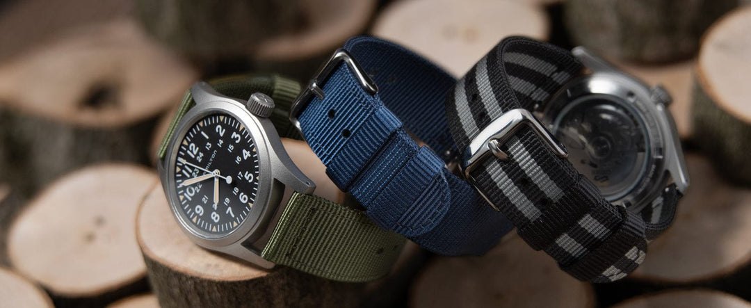 Three watches with nylon straps in olive, navy, and black-gray displayed on wooden logs