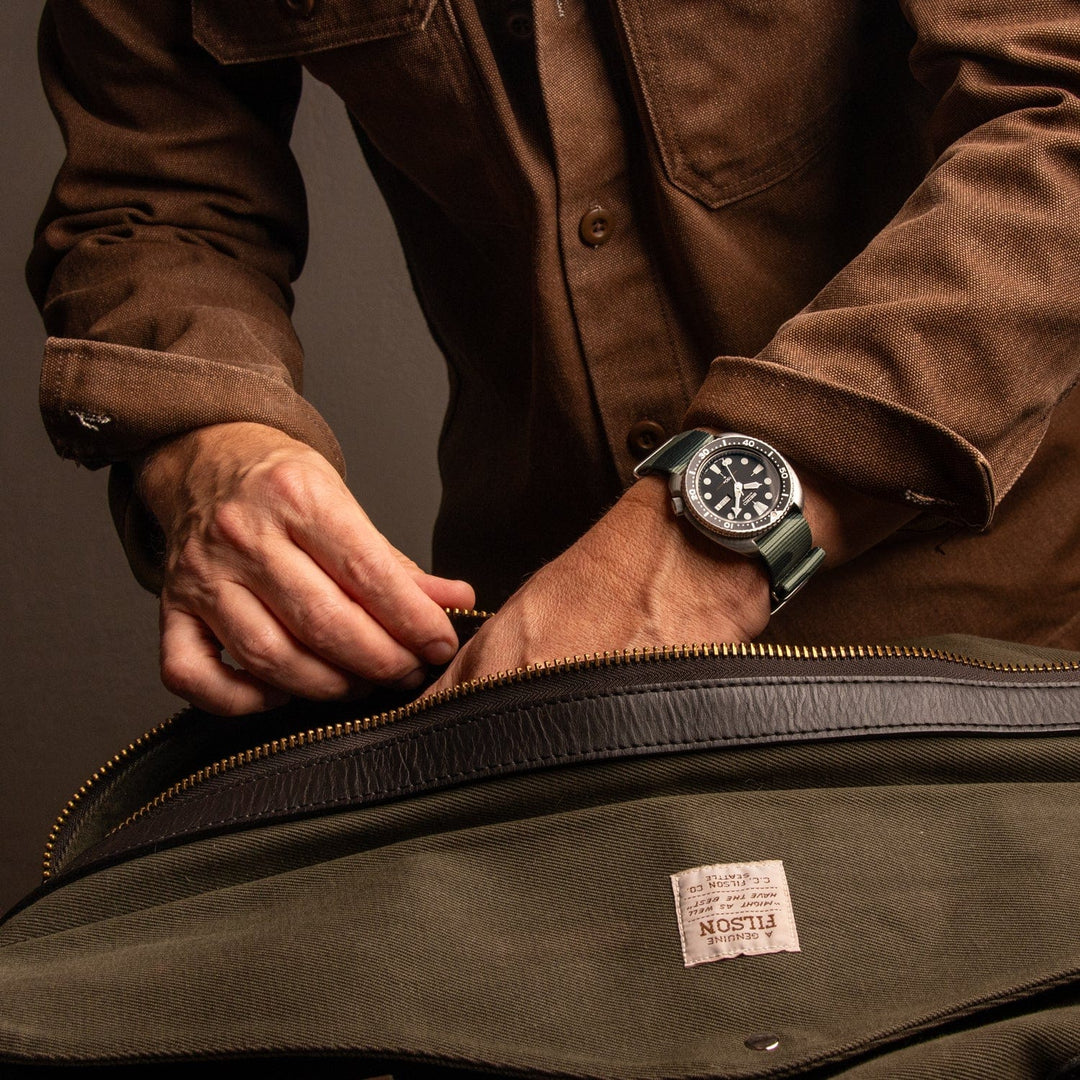 Person wearing a watch with a camouflage green military style strap while unzipping a bag
